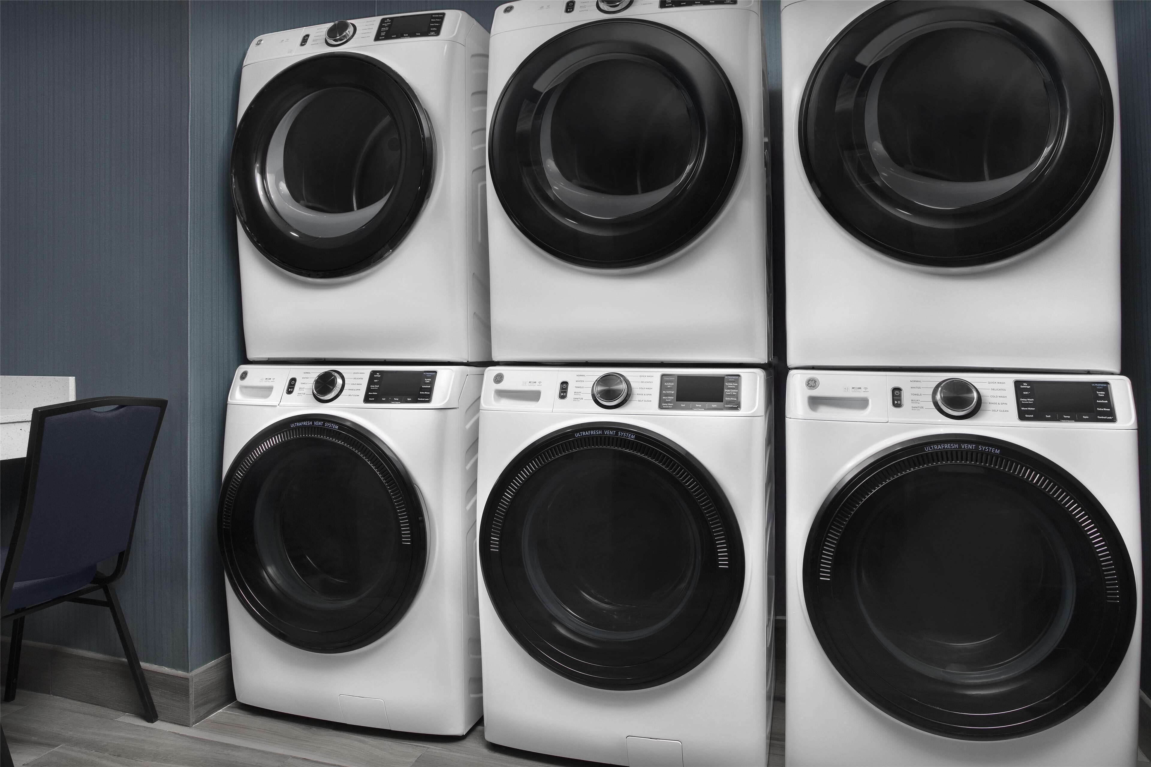 A row of four stacked front-loading washing machines in a room with a blue wall and a wooden floor.