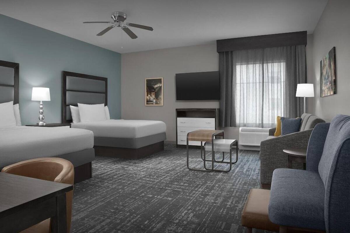 Ein Hotelzimmer mit einem Bett, zwei Nachttischen mit Lampen, einem Flachbildfernseher, einem Sofa, einem Sessel und einem Deckenventilator.