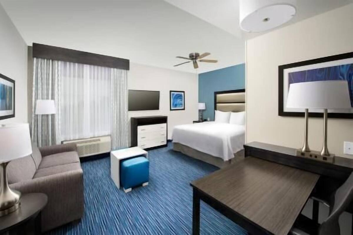 Ein Hotelzimmer mit einem Bett, einem Schreibtisch, einem Stuhl, einem kleinen Tisch, einer Couch, einem Deckenventilator und einem Fenster mit Vorhängen.
