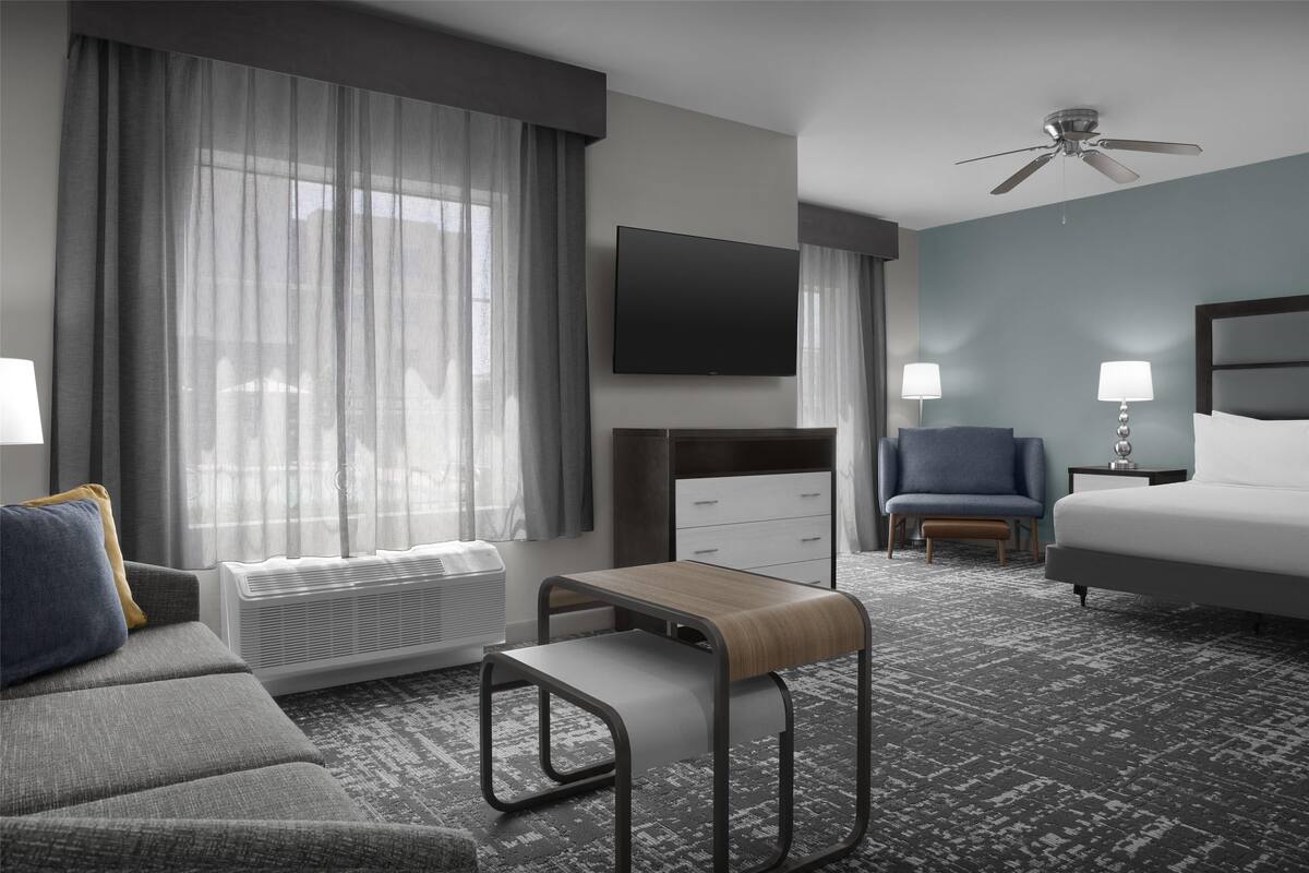 Ein Hotelzimmer mit einer Couch, einem kleinen Tisch, einem Bett, einem Fernseher, einem Deckenventilator und einem Fenster mit Vorhängen.