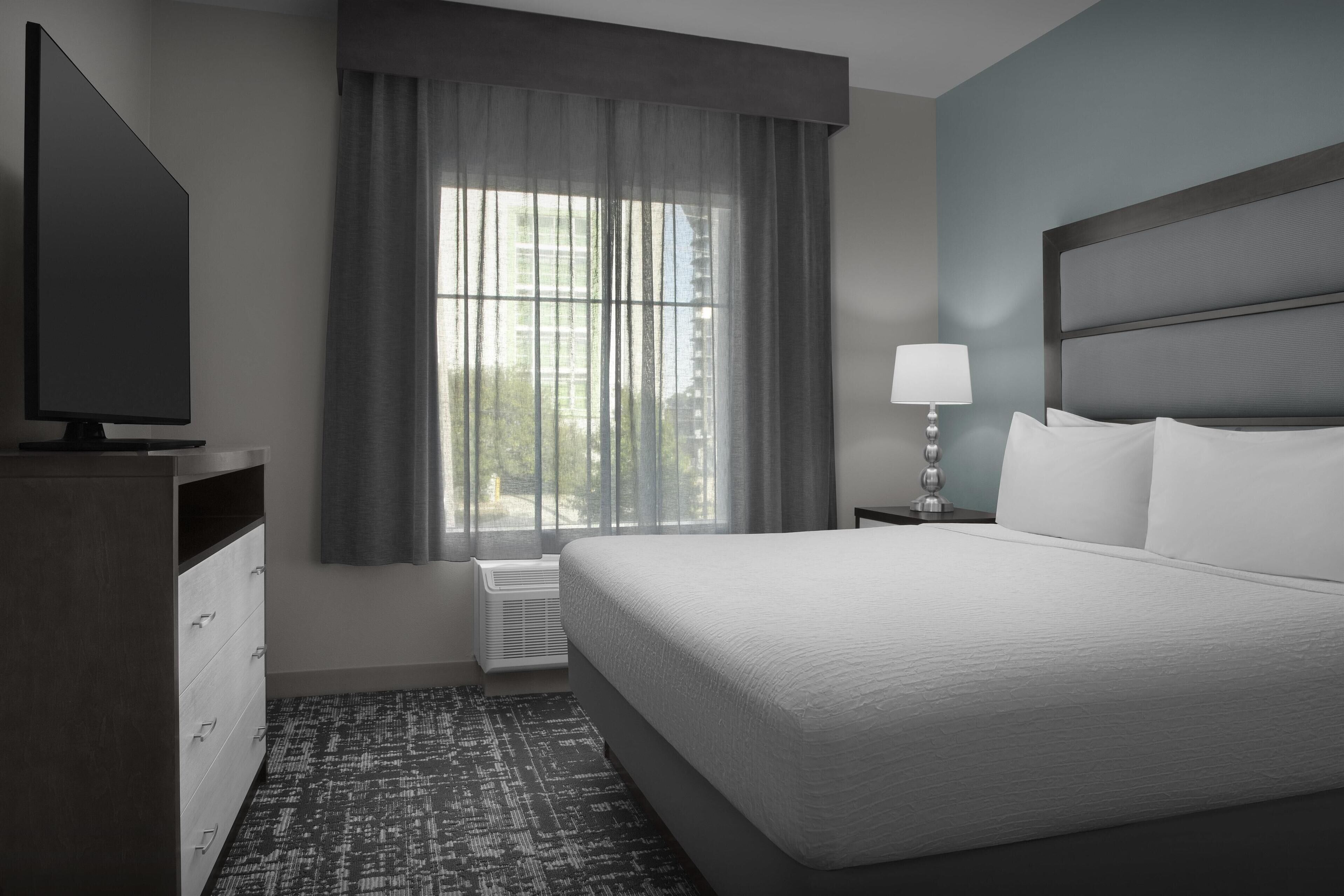 A hotel room with a large bed, a TV, a nightstand with a lamp, and a window with curtains.
