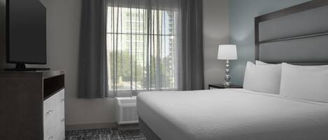 Habitación de hotel con una cama grande, un televisor, una mesita de noche con lámpara y una ventana con cortinas.