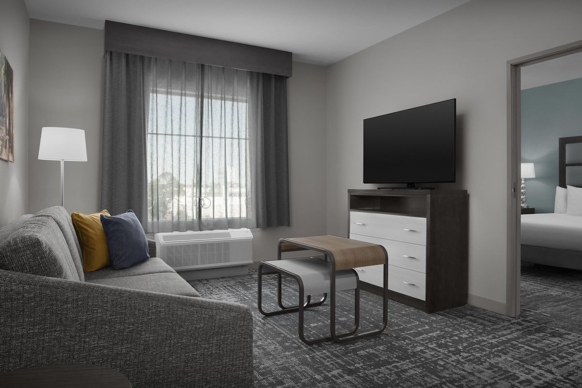 Ein modernes Hotelzimmer mit grauem Sofa, einem kleinen Holztisch, einem Flachbildfernseher und einem großen Fenster mit grauen Vorhängen.