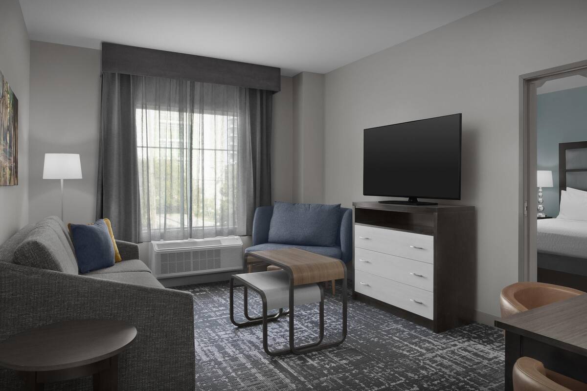 Ein modernes Hotelzimmer mit grauem Sofa, einem Holznachttisch, einem Flachbildfernseher, einem Schreibtisch mit Stuhl und einem großen Fenster mit Vorhängen.