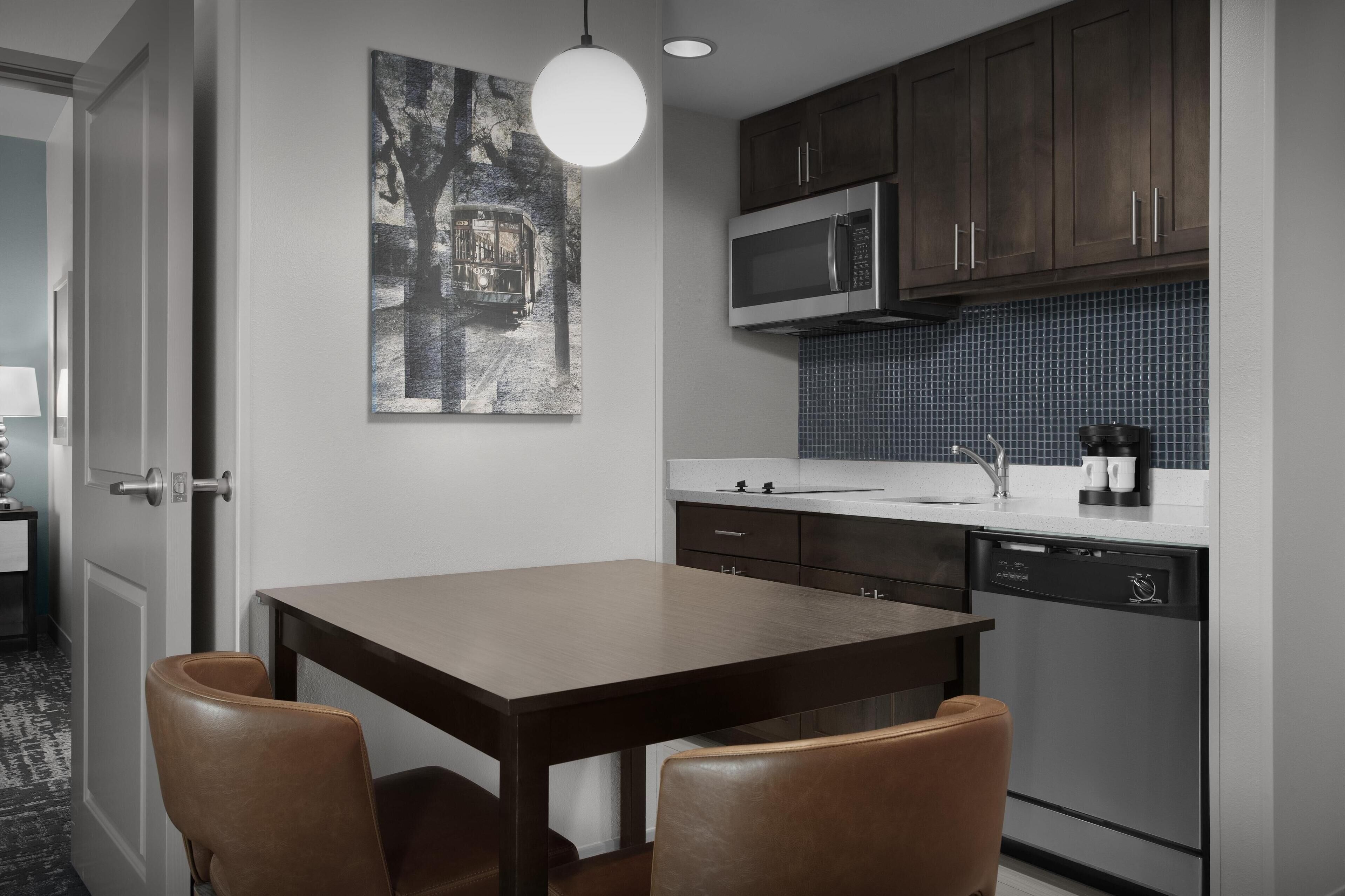 A modern kitchen with dark wooden cabinets, a stainless steel microwave, and a dishwasher. There is a dining table with two chairs and a pendant light above it.