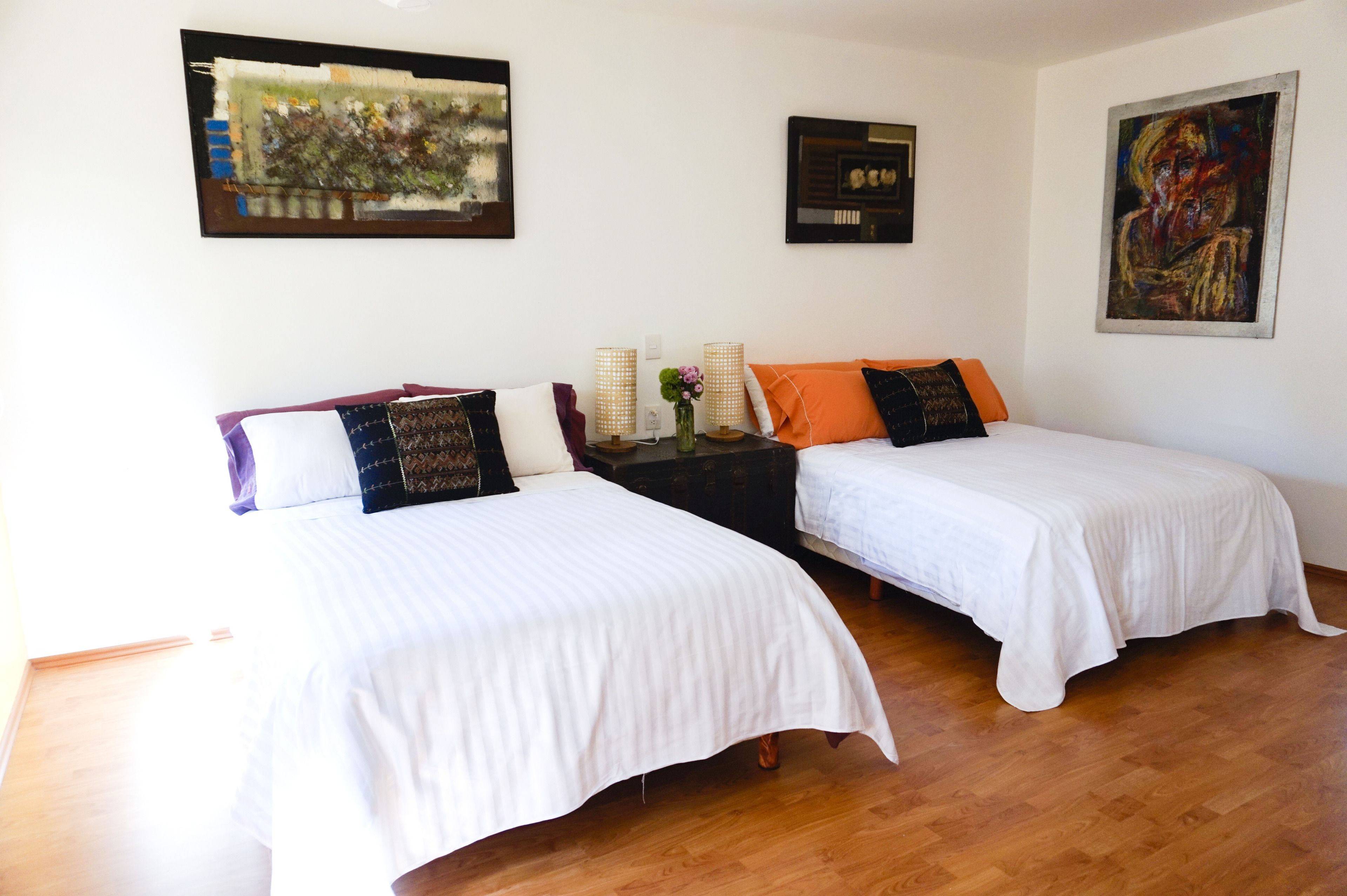 Two single beds with white linens, a nightstand with a vase and two lamps, and artwork on the walls.