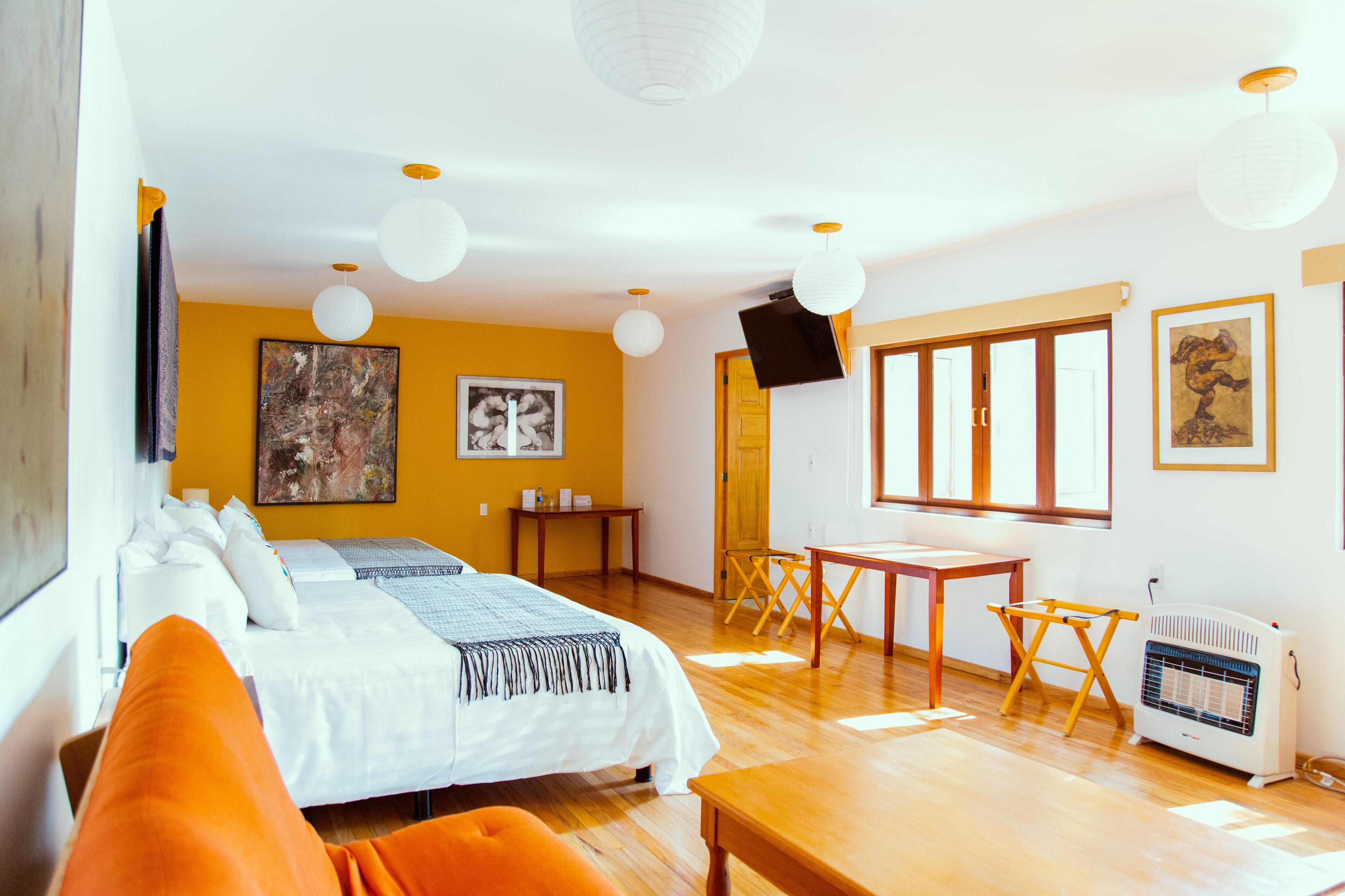 A hotel room with a bed, orange pillows, a desk, a chair, a heater, and artwork on the walls.