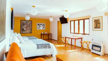 A hotel room with a bed, orange pillows, a desk, a chair, a heater, and artwork on the walls.