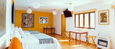 A hotel room with a bed, orange pillows, a desk, a chair, a heater, and artwork on the walls.