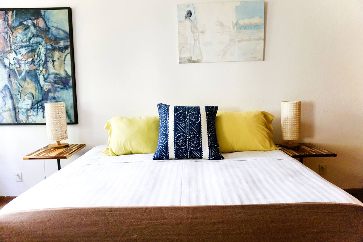 A neatly made bed with a white comforter and two pillows, one yellow and one with a blue pattern, flanked by two bedside lamps on wooden nightstands.