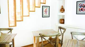 A dining area with wooden tables and chairs, framed artwork on the walls, and a white shelf with decorative items.