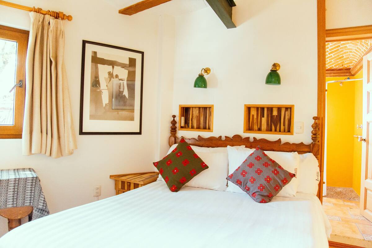 A bedroom with a wooden bed, two pillows, a framed picture, and a small table.