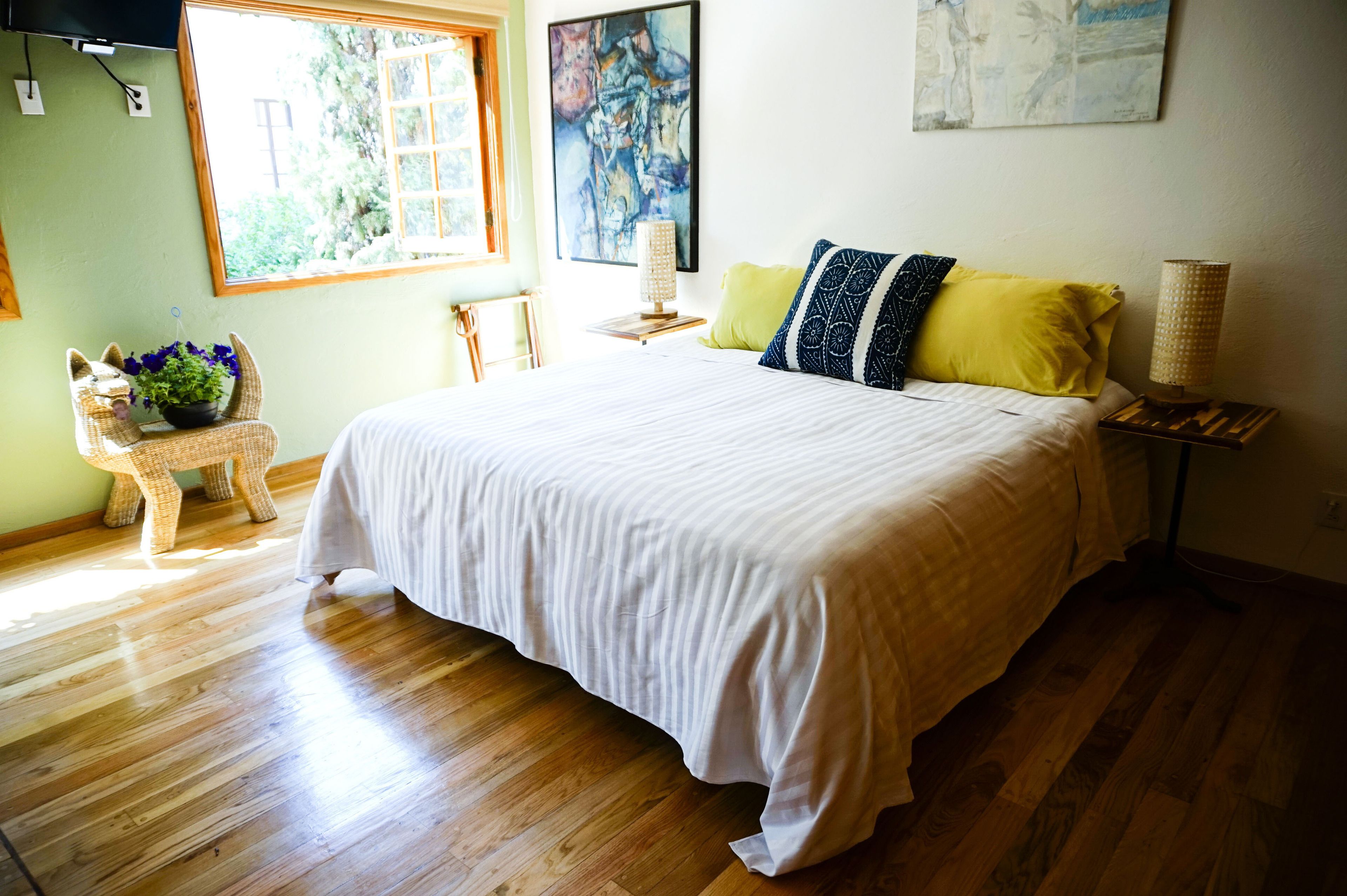 A bedroom with a large bed, a window, a painting, a lamp, and a wicker chair with a plant.