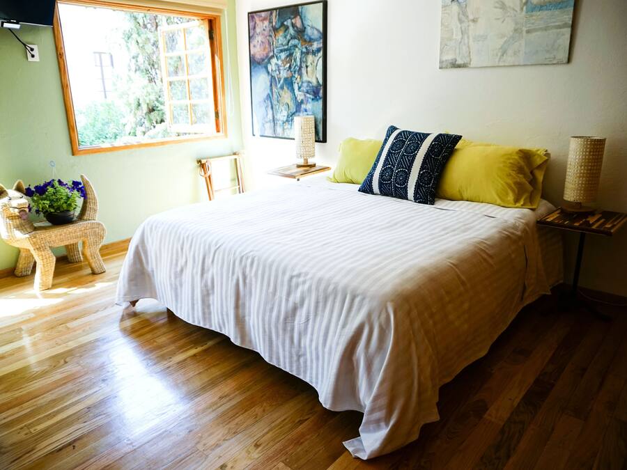 A bedroom with a large bed, a window, a painting, a lamp, and a wicker chair with a plant.