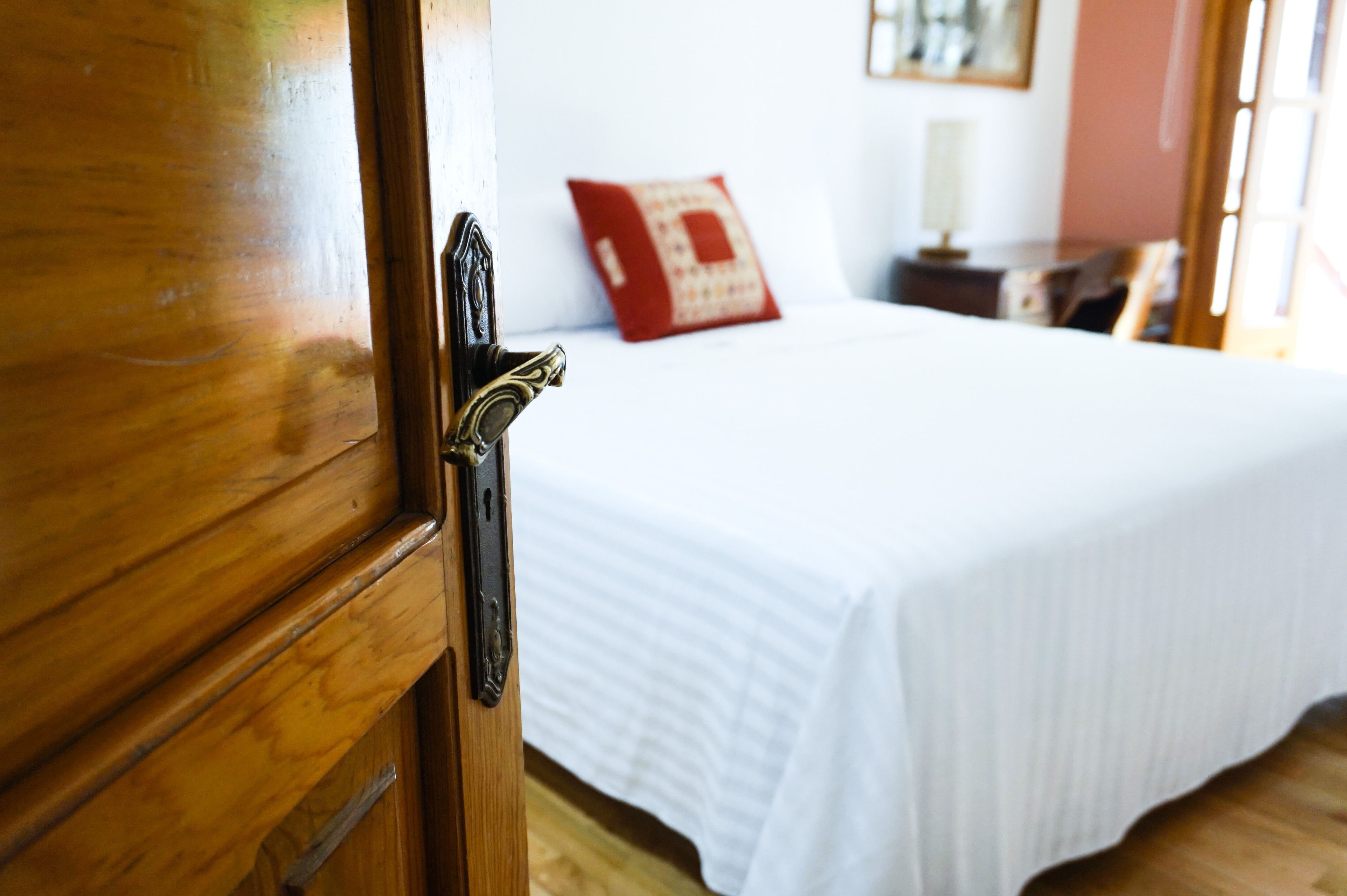 A wooden door with a brass handle, a white bed with a red pillow, and a bedside table with a lamp.
