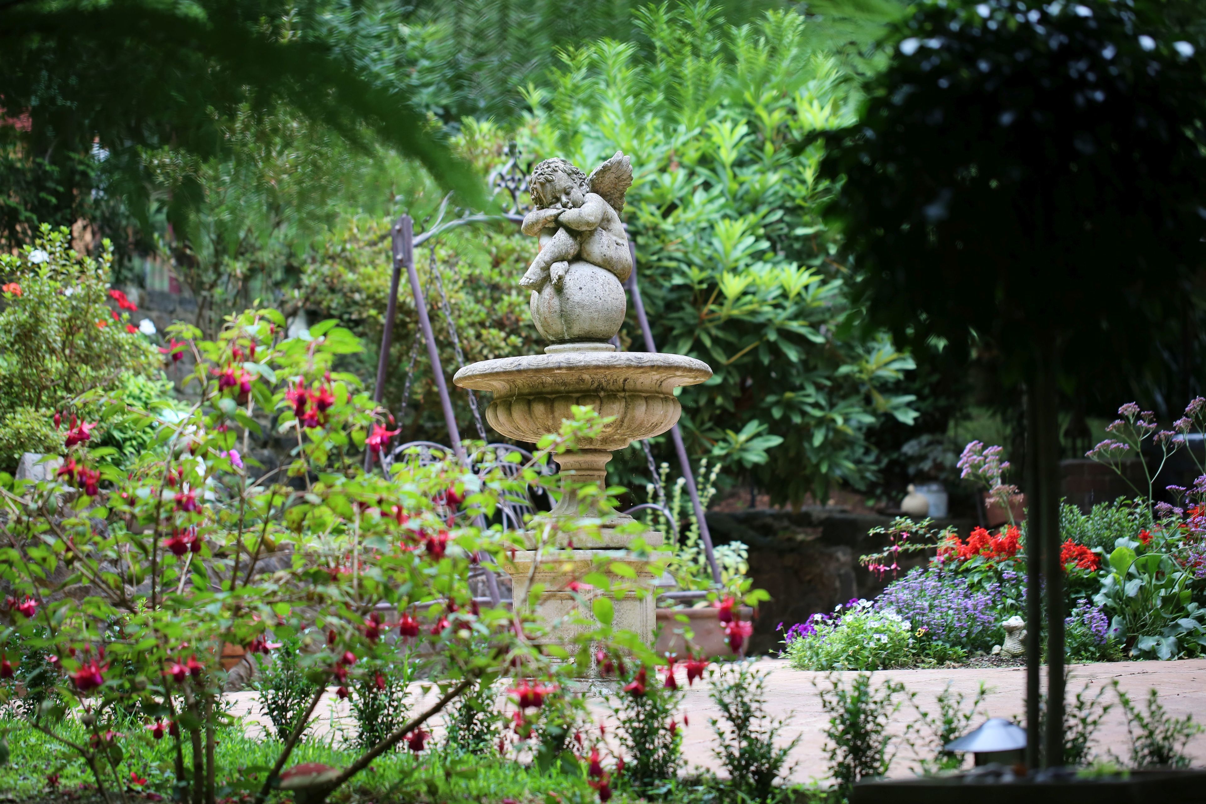 Un jardin agrémenté d’une fontaine en pierre surmontée d’une statue d’un putot, le tout entouré d’une végétation luxuriante et de plantes en fleurs.