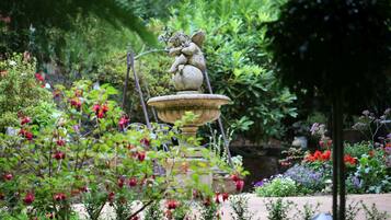 Un jardin agrémenté d’une fontaine en pierre surmontée d’une statue d’un putot, le tout entouré d’une végétation luxuriante et de plantes en fleurs.