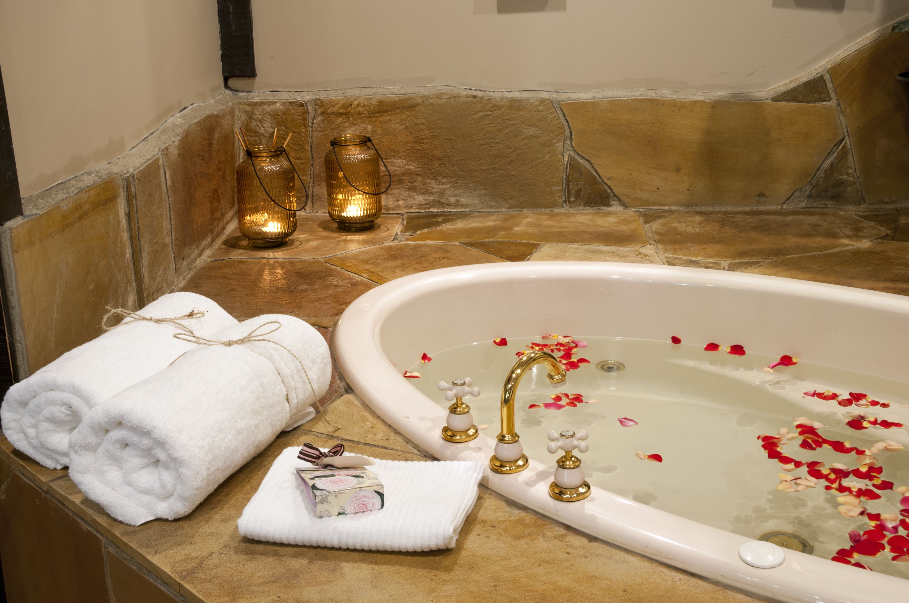 Una bañera con pétalos de rosa, una toalla y una vela sobre un suelo de baldosas de piedra.
