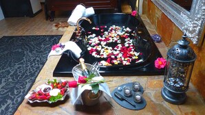 A room with a large bathtub filled with rose petals, a small table with a bottle and a rose, and a mirror with swan decorations. - Aquila Nova Retreat (The Patch)