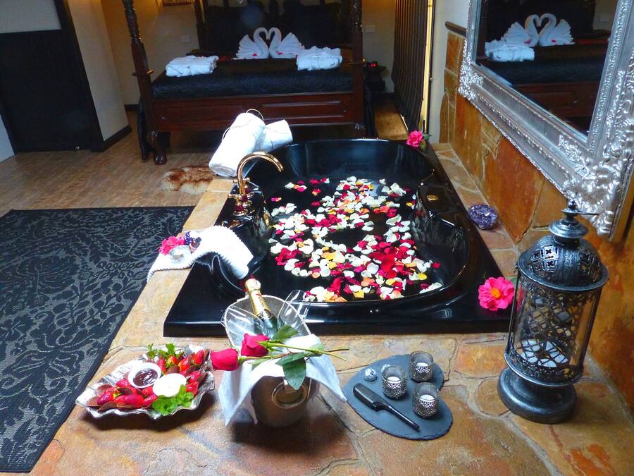 A room with a large bathtub filled with rose petals, a small table with a bottle and a rose, and a mirror with swan decorations.