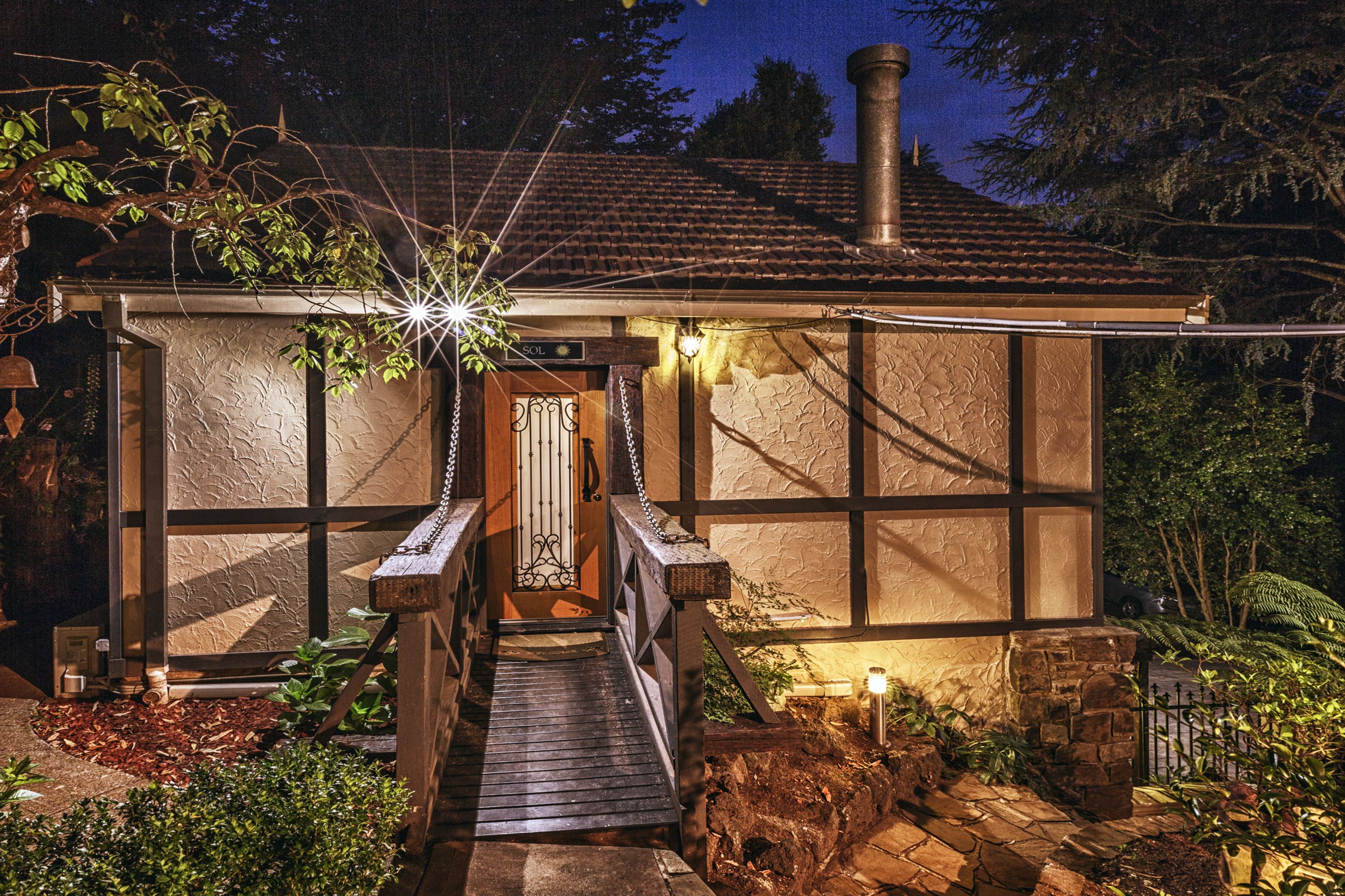 A rustic house with a wooden staircase leading to a door, surrounded by greenery and a stone pathway.