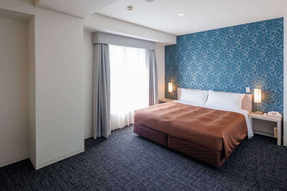 Quarto de hotel com uma cama grande, mesa de cabeceira com abajur, parede com padrão azul e janela com cortinas.