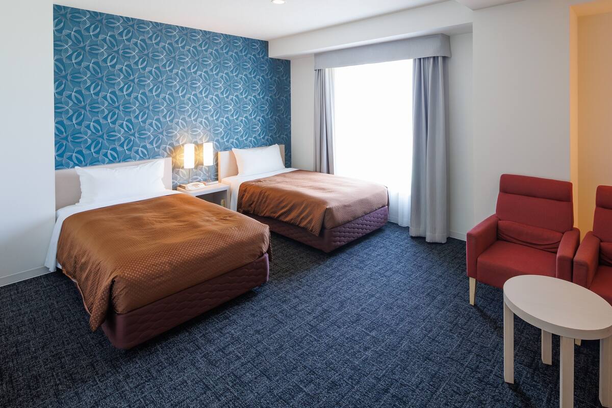 Quarto de hotel com duas camas, uma poltrona vermelha, uma mesinha e uma parede estampada.