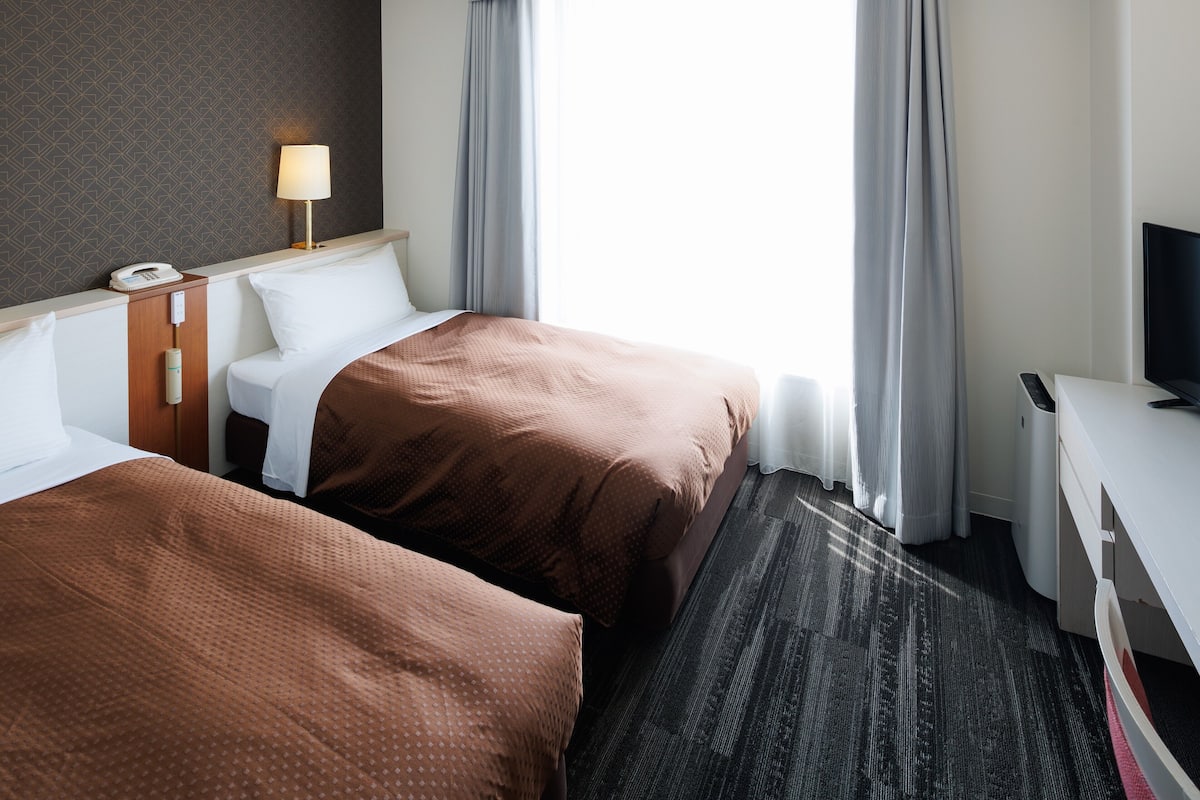 Quarto de hotel com duas camas, uma escrivaninha, uma luminária e uma televisão.
