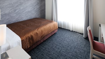 A hotel room with a bed, a chair, a desk, and a window with curtains.