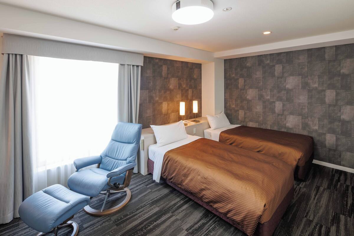 Quarto de hotel com cama, área de estar com uma poltrona, mesa de cabeceira e luminária fixada na parede.