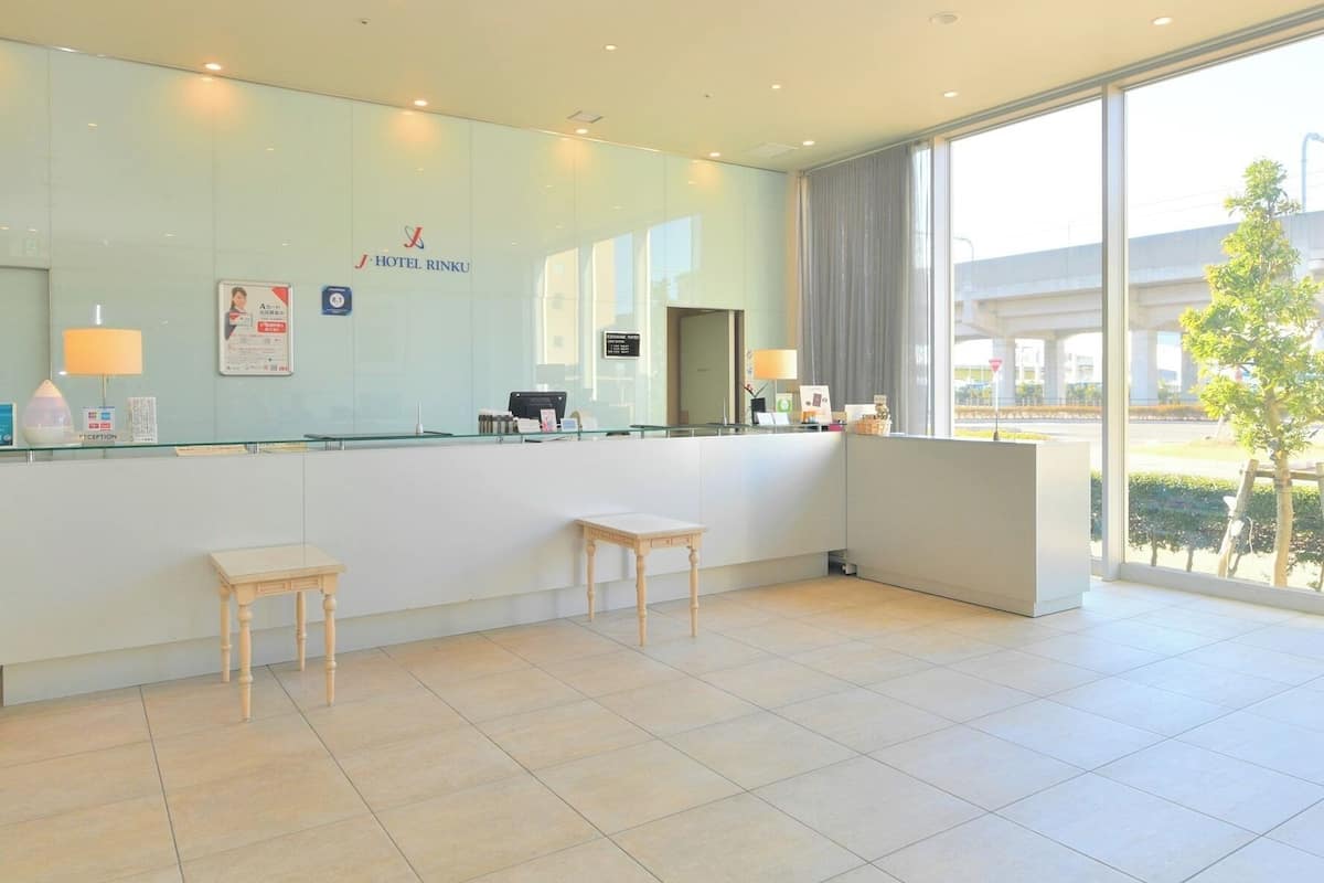Uma área de recepção de hotel com balcão de vidro, mesa de atendimento, dois taboretes de madeira e uma grande janela com vista para o exterior.