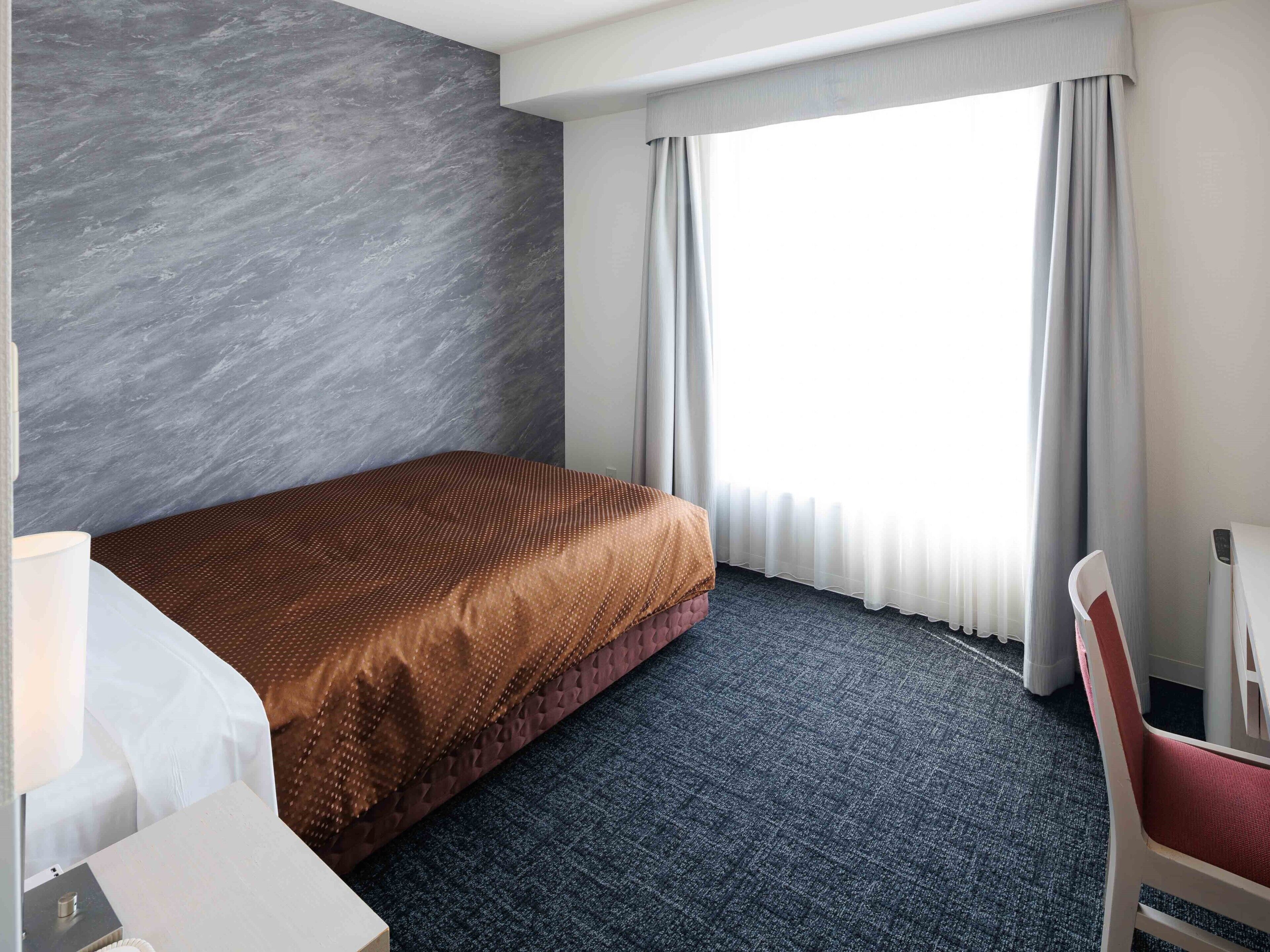 A hotel room with a bed, a chair, a desk, and a window with curtains.