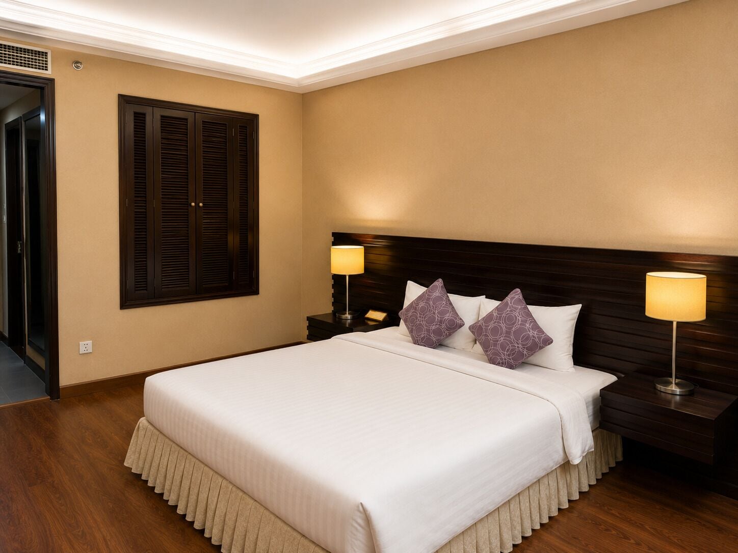 A hotel room with a large bed, two bedside lamps, a wooden headboard, and a window with dark shutters.
