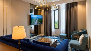 A hotel room with a blue sofa, a small table, a lamp, a TV on the wall, and a view of greenery outside. - Savan Resorts (Savannakhet)