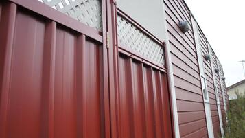 Ein roter Metallzaun mit Gitterspitze, neben einem Gebäude mit horizontaler Schalung und weißem Fensterrahmen.