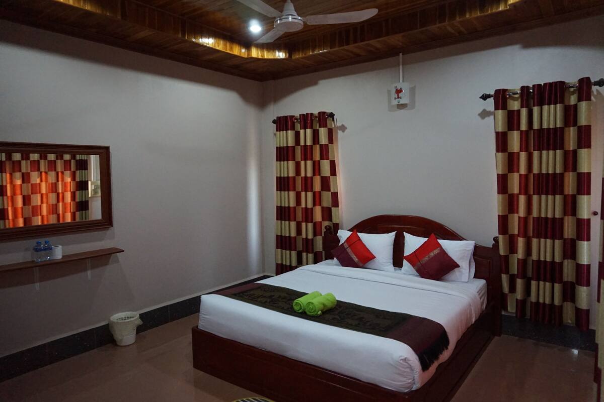 A neatly made bed with a green towel, red and beige curtains, and a ceiling fan.