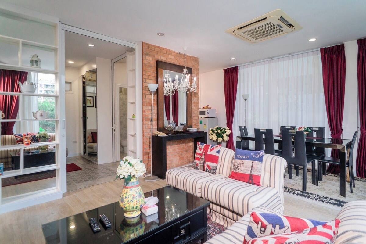 A modern living room with a sofa, a coffee table, and a dining area with a table and chairs. There is a fireplace with a mirror above it, and a chandelier hanging from the ceiling.