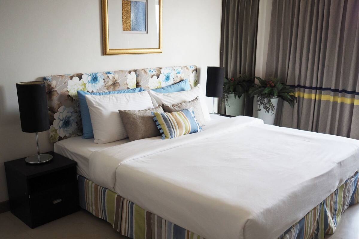 A neatly made bed with a striped bed skirt, a headboard with a floral pattern, two bedside lamps, and a framed picture on the wall.