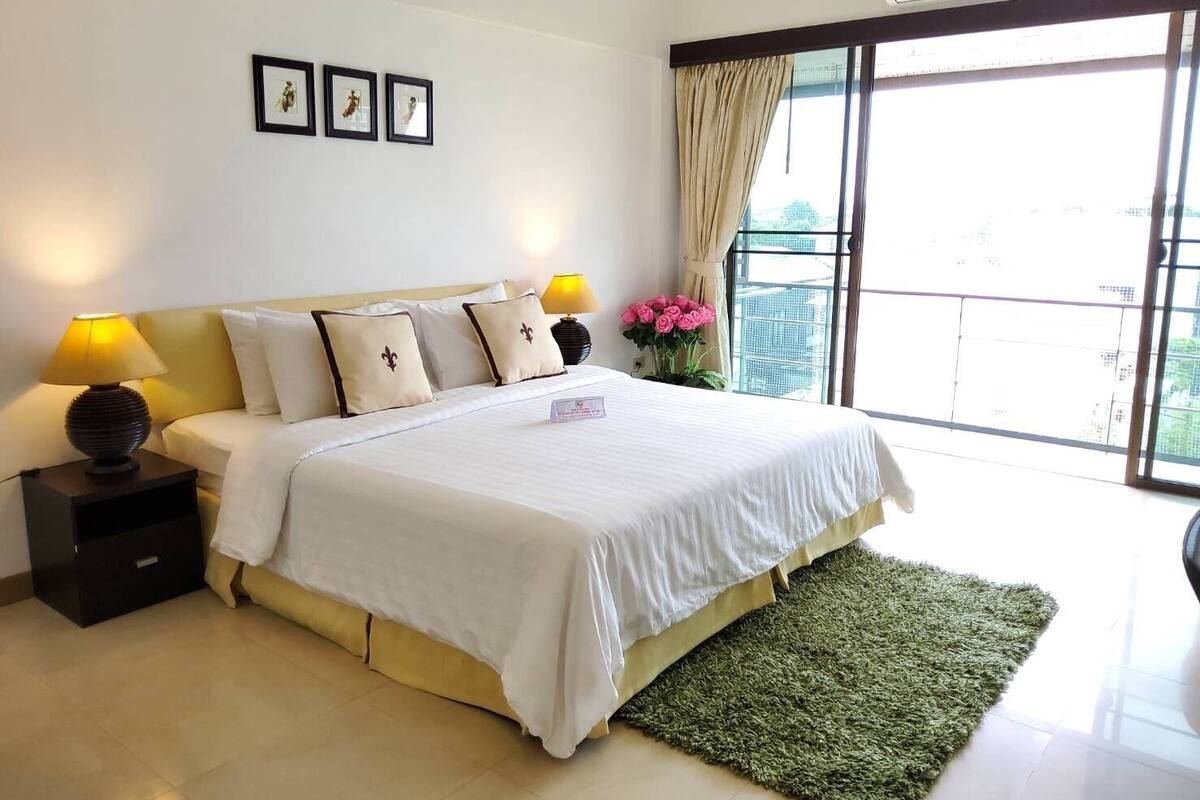 A bedroom with a bed, two bedside lamps, a nightstand, a green rug, a window with curtains, and framed pictures on the wall.