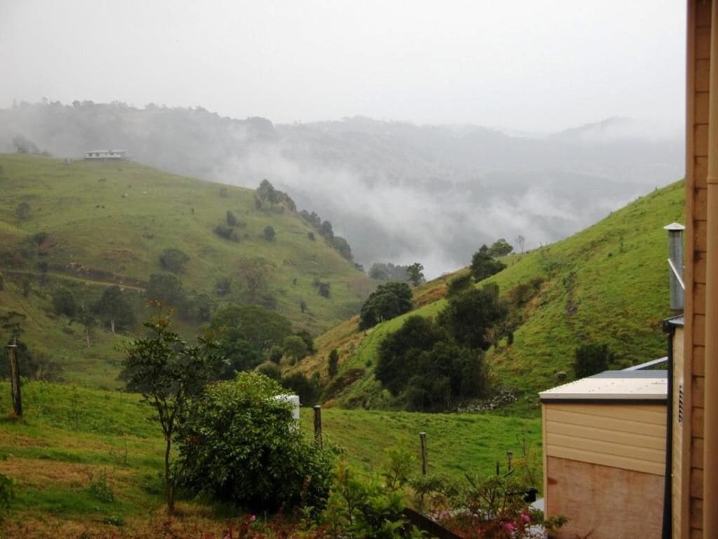 Un paysage rural avec des collines ondoyantes, un petit bâtiment et une maison éloignée, dont on ne distingue qu’une partie à travers le brouillard.