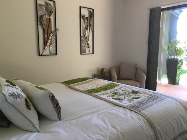 A bedroom with a bed, two framed artworks on the wall, a chair, and a view of greenery outside.
