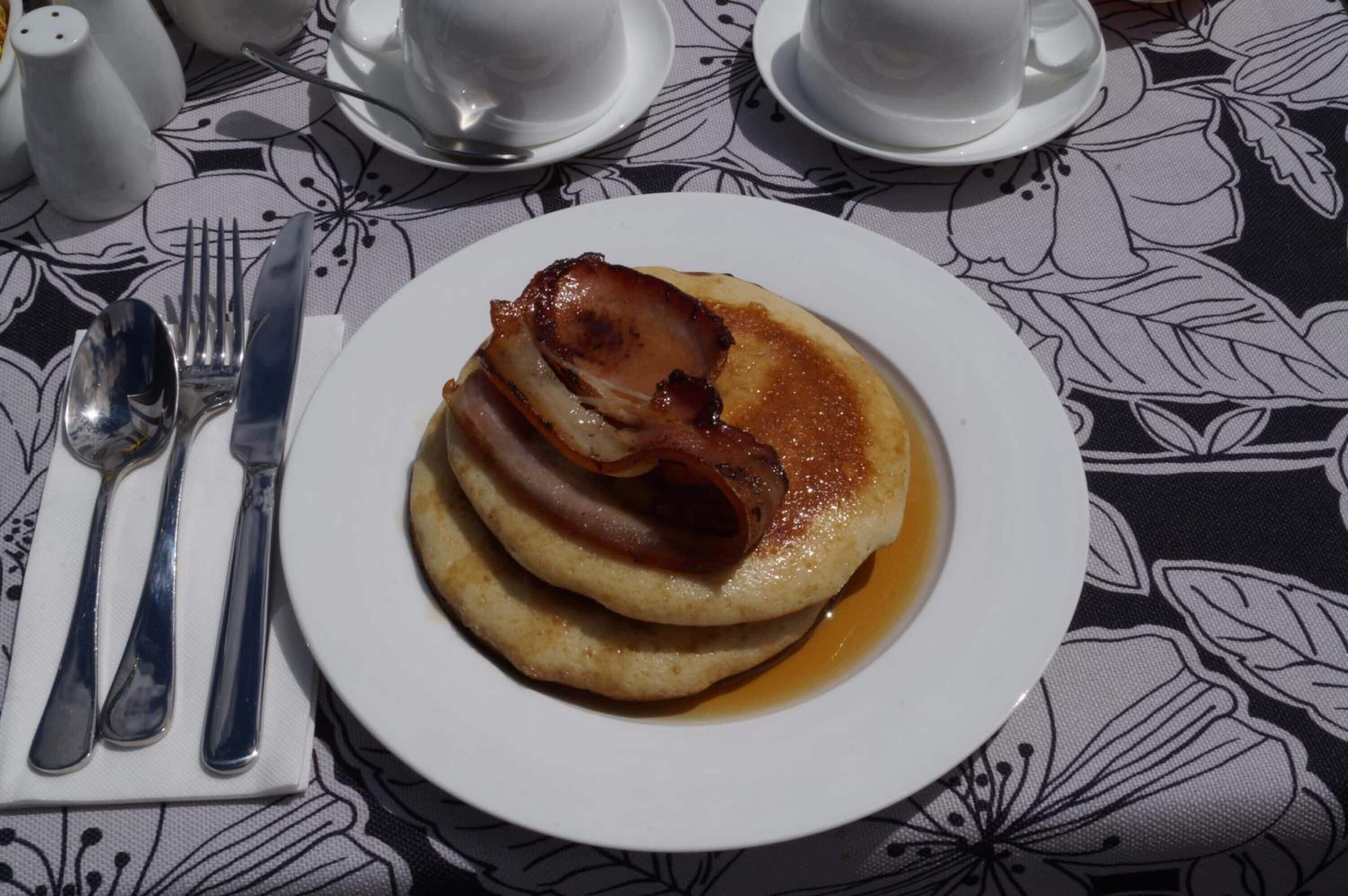 A plate of pancakes with bacon and syrup, accompanied by a fork and knife on a table with a floral tablecloth.