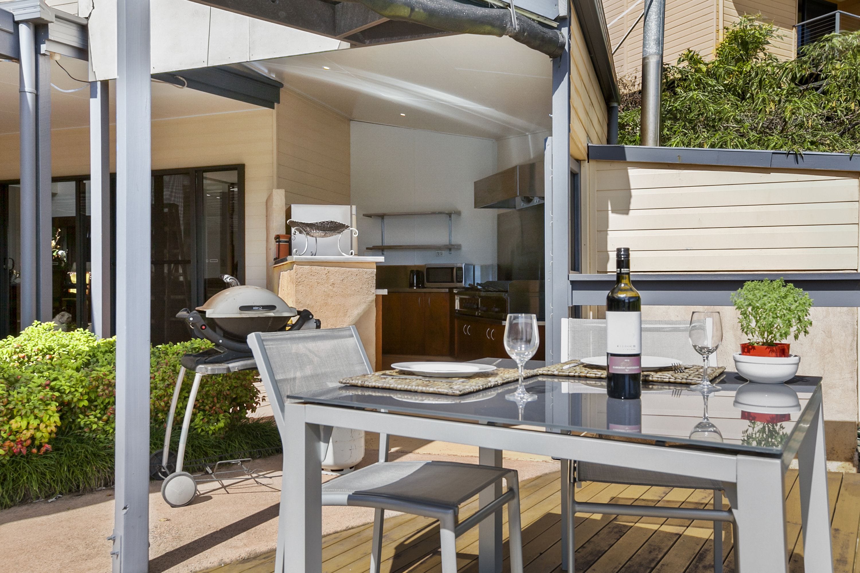 An outdoor dining area with a table set for two, a grill, and a view of the kitchen.