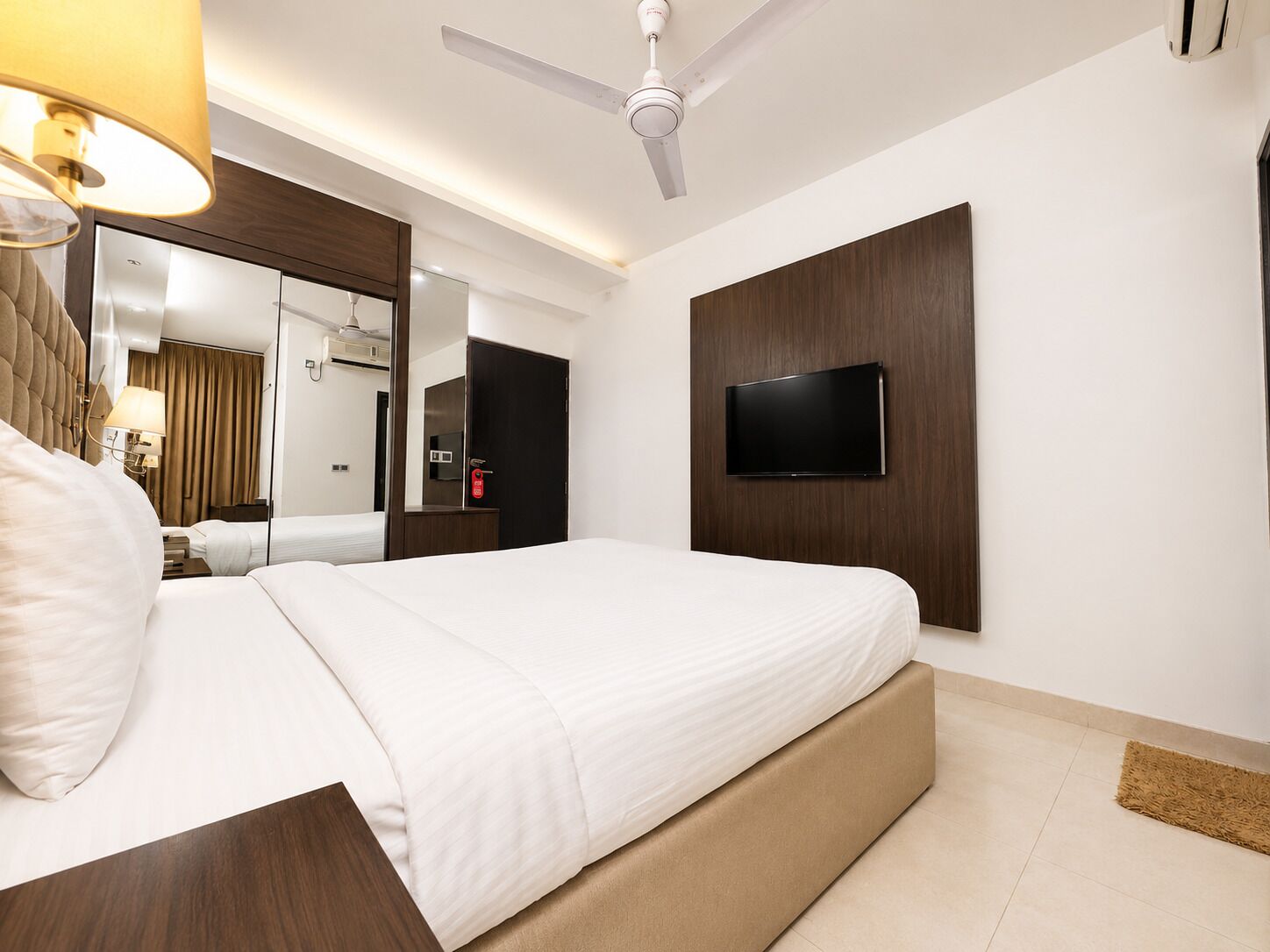 A hotel room with a bed, a flat-screen TV mounted on the wall, a ceiling fan, and a mirror.