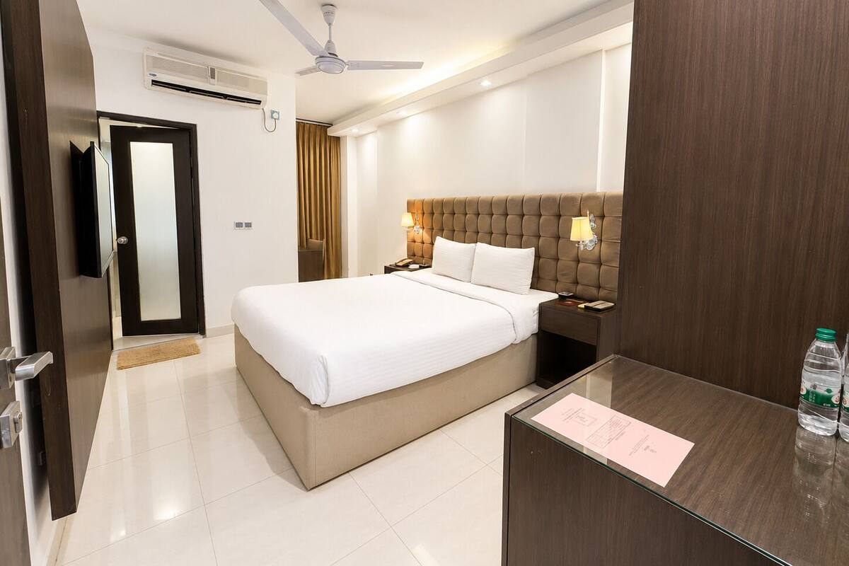 A hotel room with a bed, a desk, a chair, a TV, a ceiling fan, and a door.