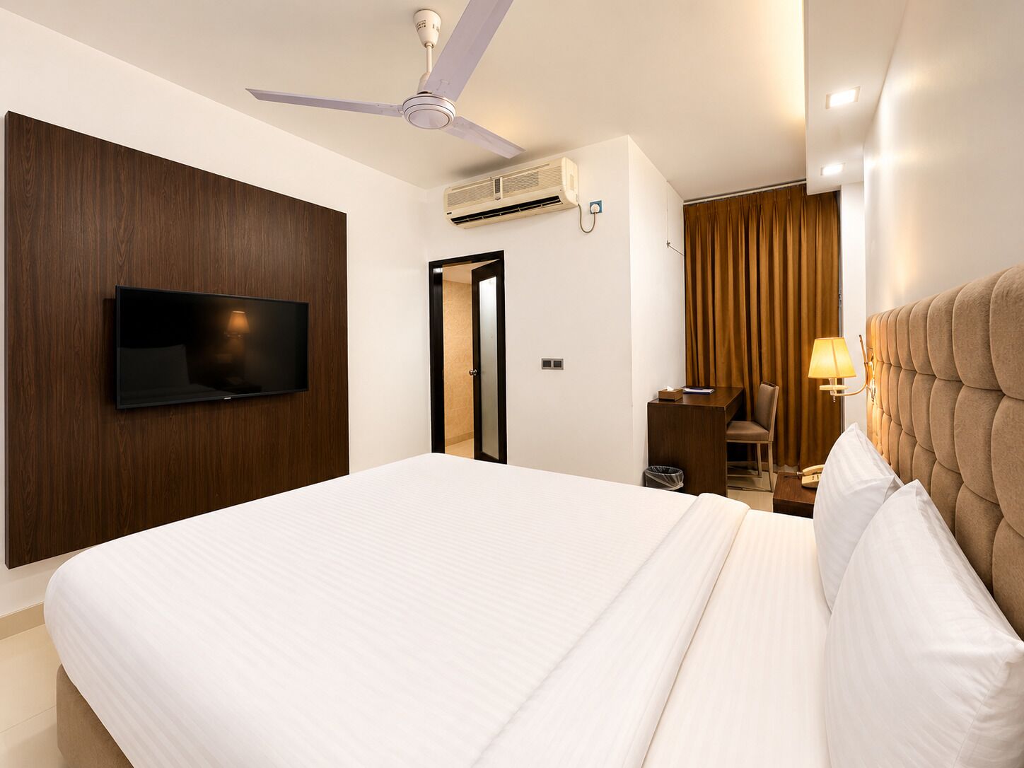 A hotel room with a bed, a TV mounted on the wall, a ceiling fan, an air conditioning unit, a desk with a lamp, and a window with curtains.