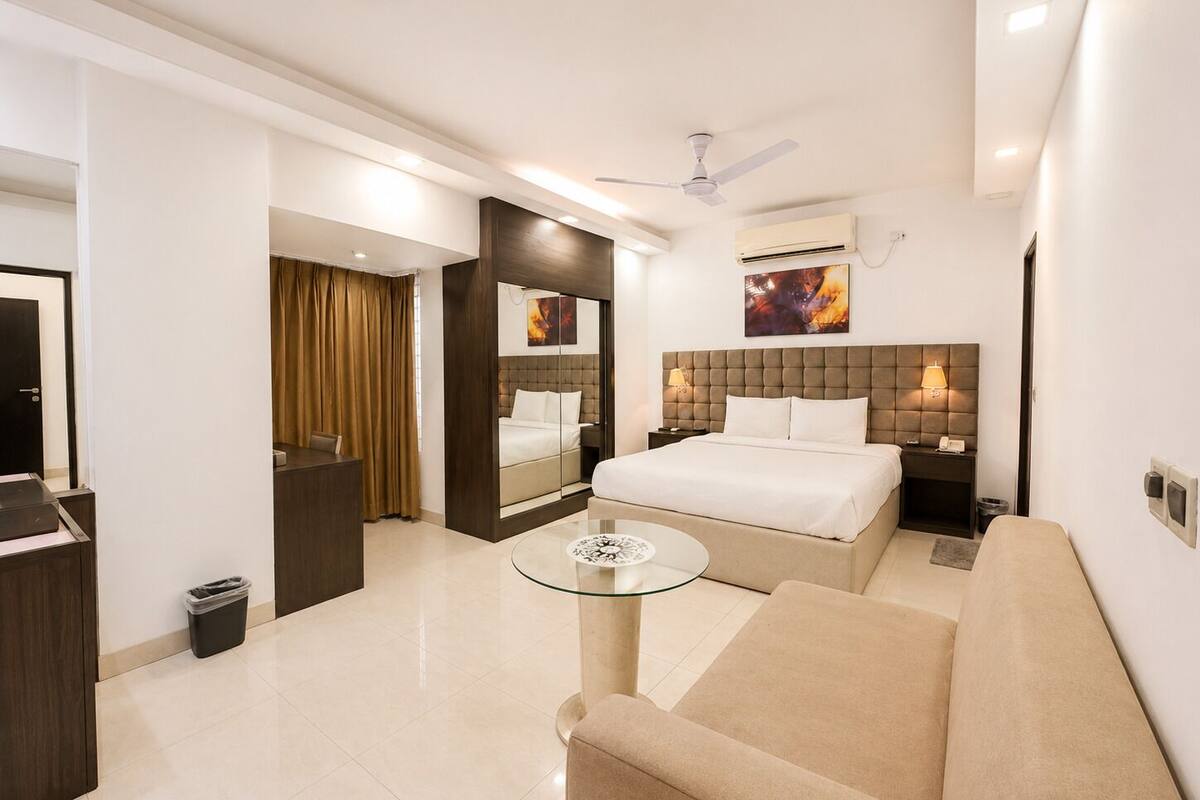 A hotel room with a bed, a sofa, a glass coffee table, a ceiling fan, and a wall-mounted air conditioning unit.