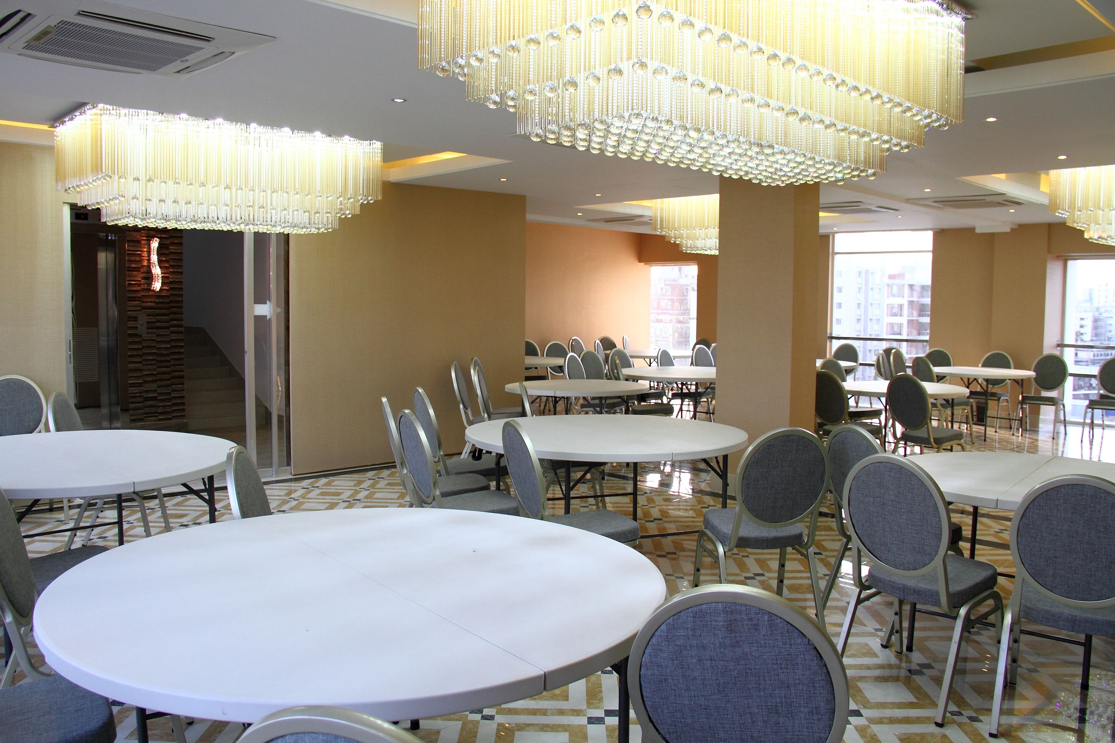 A conference room with multiple tables and chairs, large chandeliers, and a view of buildings through the windows.
