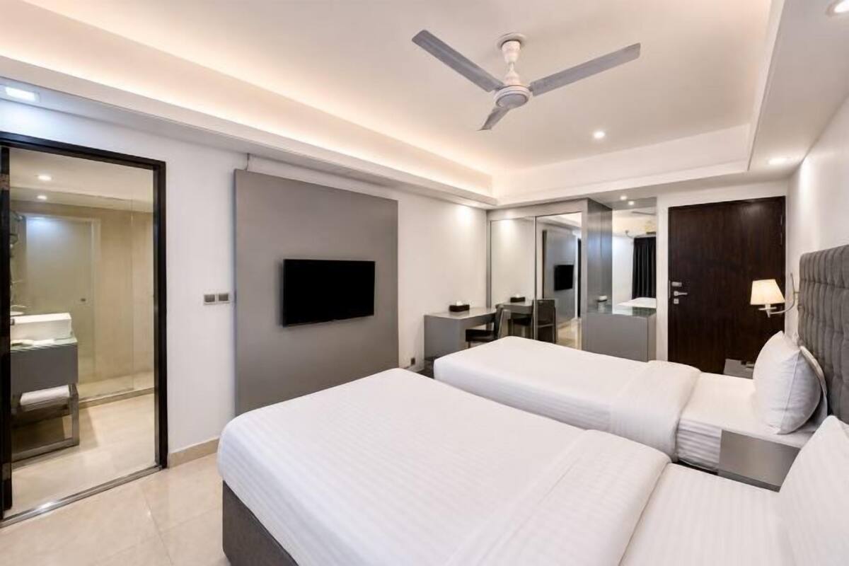 A modern hotel room with two beds, a TV, a ceiling fan, and a bathroom visible through an open door.