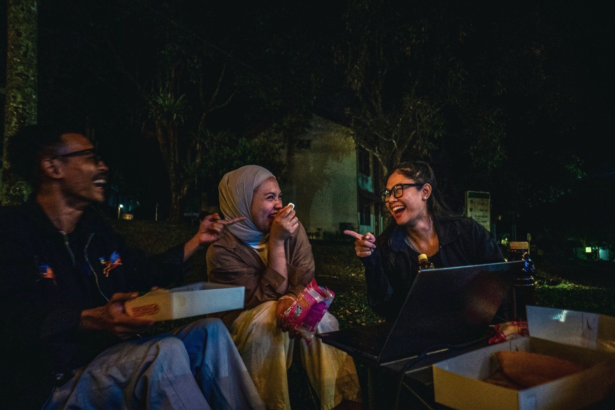 Three people sitting outdoors at night with pizza boxes, a laptop, and a smartphone.